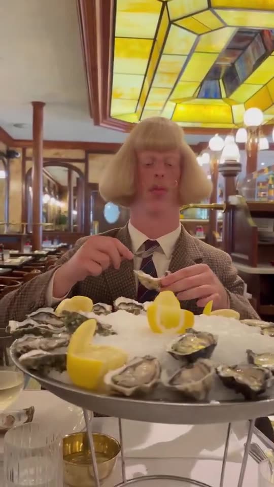 Jean Phil, an AI character with a blonde bob and a houndstooth jacket, eating oysters in a brasserie under a stained-glass ceiling